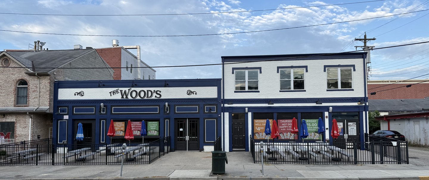 The two storefronts at 17 N Poplar Street seen from across the road: The Wood's navy building with its name on a white sign band on the left, and the white and navy Sidebar building on the right, each with picnic tables behind a rail.