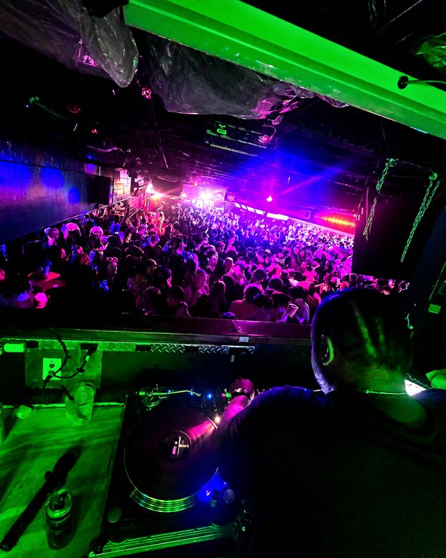 The DJ booth at The Woods, with the room filled beyond it under green and magenta light.